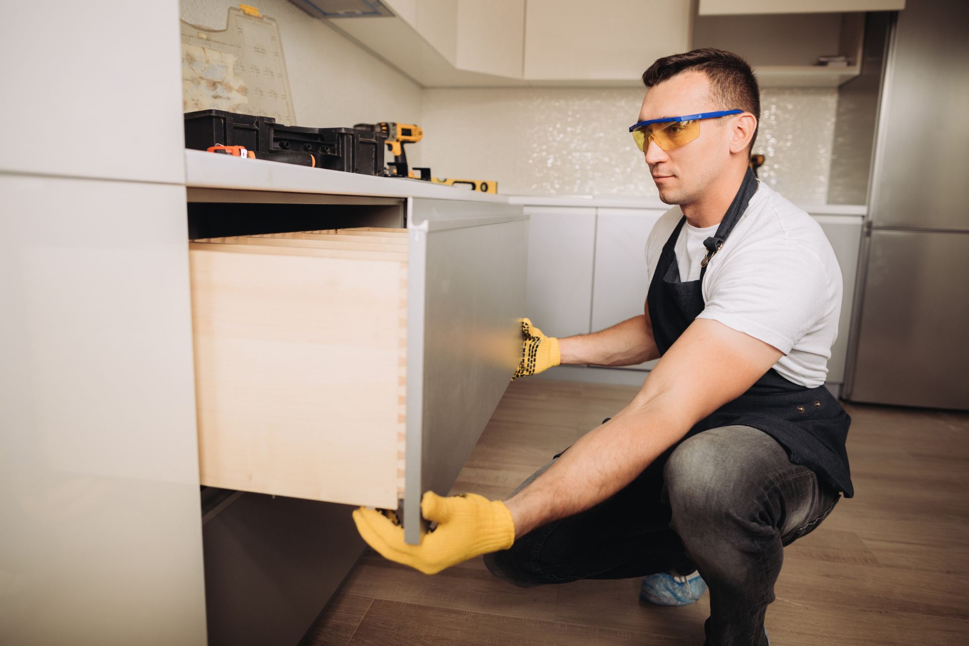 A man wearing an apron and glasses repairs a wooden cabinet, focused on his task with tools nearby.